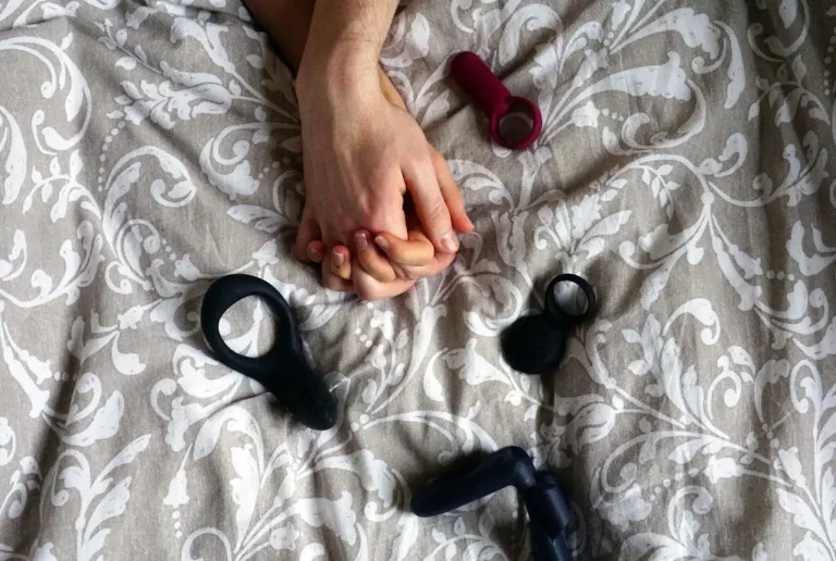 Interlaced hands on patterned bedspread surrounded by cock rings and a prostate massager.