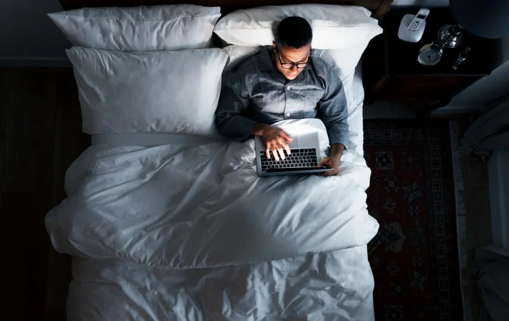 Man browsing on a laptop in bed at night, shopping privately from home
