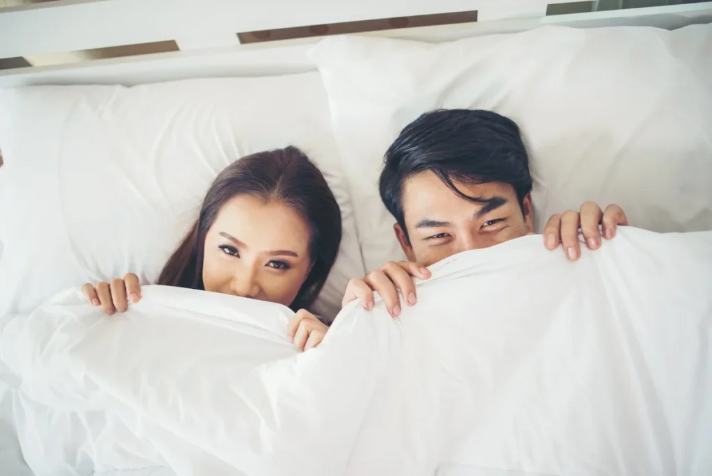 Couple peeking out from under a white blanket in bed, a playful moment of intimacy
