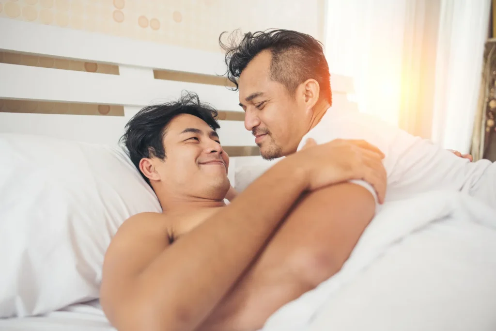 Two men holding each other in bed and smiling, the closeness foreplay is meant to build